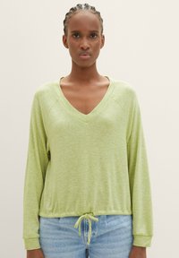 Frau mit geflochtenem Haar, die ein hellgrünes, langärmliges Oberteil mit V-Ausschnitt und Kordelzugsaum sowie helle Jeans trägt, steht vor einem einfarbigen Hintergrund.