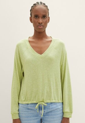 Woman with braided hair wearing a light green V-neck long-sleeve top with drawstring hem and light blue jeans, standing against a plain background.
