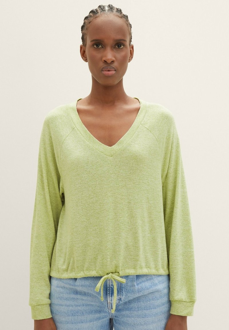 Frau mit geflochtenem Haar, die ein hellgrünes, langärmliges Oberteil mit V-Ausschnitt und Kordelzugsaum sowie helle Jeans trägt, steht vor einem einfarbigen Hintergrund.