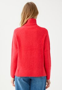 Przesadnie duży sweter z golfem w jaskrawym czerwonym kolorze, wykonany z miękkiej, teksturowanej tkaniny. Żebrowany kołnierz i dół, z opuszczonymi szwami ramion.