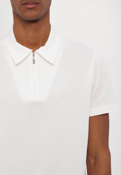 Balta polo marškinėliai su užtrauktuku ant apykaklės, trumpomis rankovėmis ir tekstūruotu audiniu, pabrėžiantys švarų ir modernų dizainą.