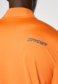 Oranges Sporthemd mit glatter Textur und Kragen; verfügt über ein dunkelgraues "SPYDER"-Logo auf dem oberen Rücken.