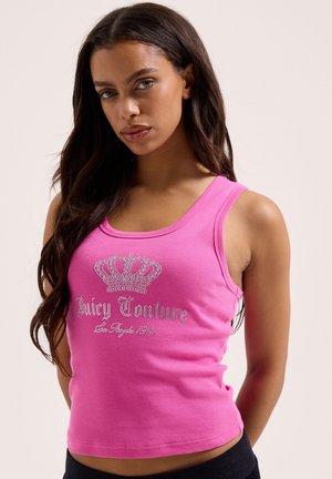 Frau mit langen dunklen Haaren, die ein knallrosa Tanktop mit einer Strasskrone und dem Schriftzug "Juicy Couture" trägt und vor einem hellen Hintergrund steht.