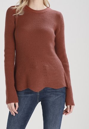 Vrouw met een roestkleurige geribbelde gebreide trui met een geronde zoom en een blauwe spijkerbroek, staand tegen een effen lichte achtergrond.