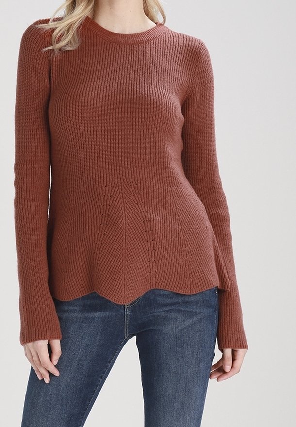 Femme portant un pull en maille côtelée couleur rouille avec un ourlet festonné et un jean bleu, debout devant un fond clair uni.