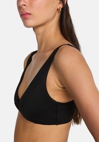 Vrouw met een zwart mouwloos topje met dunne bandjes, haar profiel zichtbaar van schouder tot bovenkant borst, met gouden oorringen.