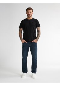 Camiseta de algodón negra con cuello redondo, combinada con jeans azul oscuro y zapatillas blancas. Presenta tatuajes visibles en los brazos.