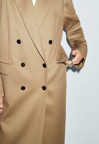 Beige Doppelreiher-Mantel mit großen schwarzen Knöpfen, strukturierten Revers und Fronttaschen. Aus strukturiertem Stoff gefertigt, der eine präzise Schneiderkunst zeigt.