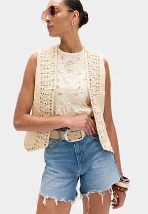 Kvinde iført broderet cremet ærmeløs vest, blondekantet bluse, denimshorts, solbriller og armbånd, poserer med hånden på hoften.