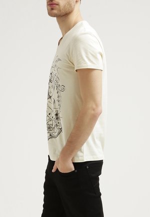 Camiseta de algodón color crema con mangas cortas y escote en V, con un diseño gráfico impreso en negro en el frente. Llevada con pantalones negros.