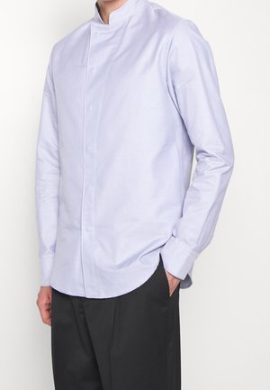 Personne portant une chemise violette claire à manches longues avec col mao et un pantalon noir, une main dans la poche, debout devant un fond blanc.