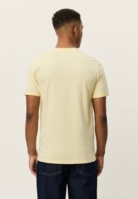 Κίτρινο βαμβακερό t-shirt με κοντά μανίκια, στρογγυλή λαιμόκοψη και απαλό ύφασμα. Η πίσω πλευρά εμφανίζει ένα απλό σχέδιο χωρίς διακοσμήσεις.