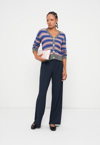 Flerfarget stripet cardigan med grå detaljer, knappelukking, kombinert med marineblå stripete bukser og en rosa clutch. Sorte leiligheter fullfører antrekket.