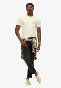 Camiseta de algodón color beige con estampado "SUPER DRY", jeans oscuros y una camisa de franela a cuadros atada a la cintura. Zapatillas de colores claros completan el atuendo.