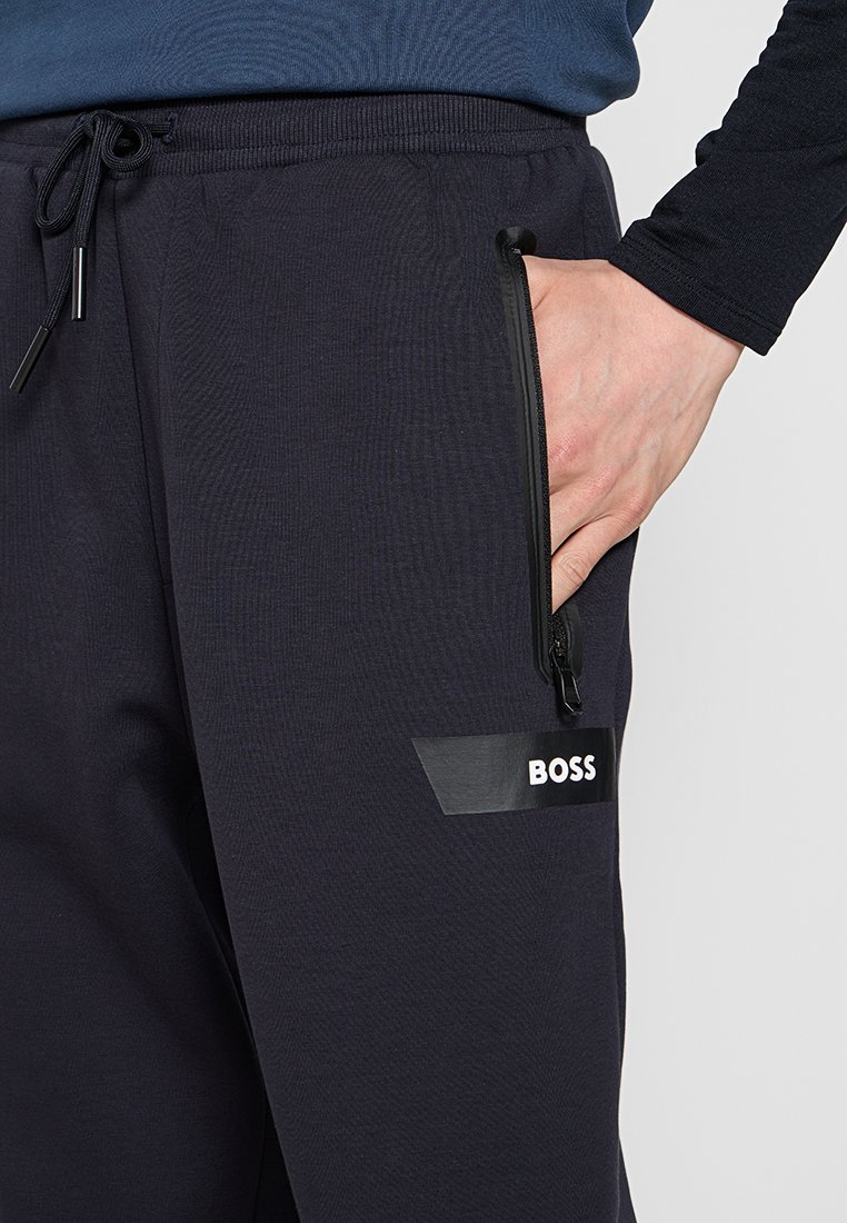 Hand in de met ritssluiting opgeborgen zak van zwarte BOSS trainingsbroek met trekkoordtailleband, gedragen met een donker langemouwenshirt.