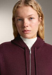 Mladý človek so svetlou pokožkou, blond vlasmi a modrými očami, oblečený v bielom roláku a fialovej mikine na zips, pozerajúci sa dopredu.