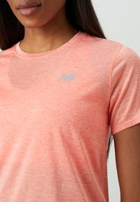 Κοντινή λήψη ενός ατόμου που φοράει ένα κοραλί αθλητικό μπλουζάκι με το λογότυπο της New Balance σε ανοιχτό γκρι στο στήθος.