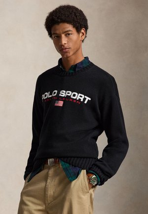 Młody mężczyzna w czarnym swetrze Polo Sport Ralph Lauren, zielonej koszuli w kratę, beżowych spodniach i zielonym zegarku na nadgarstku na neutralnym tle.