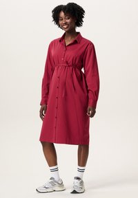 Rotes, tailliertes Umstandskleid mit Gürteldetails, langen Ärmeln und knielangem Saum. Getragen mit weißen Socken und grauen Turnschuhen.