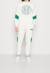 Felpa bianca con accenti verdi e logo sul davanti, abbinata a pantaloni della tuta bianchi con toppe verdi e logo. Sneakers bianche.