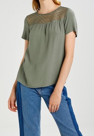 Femme portant un chemisier vert olive à manches courtes avec un détail en dentelle sur l'empiècement et un jean bleu bicolore, debout devant un fond uni.