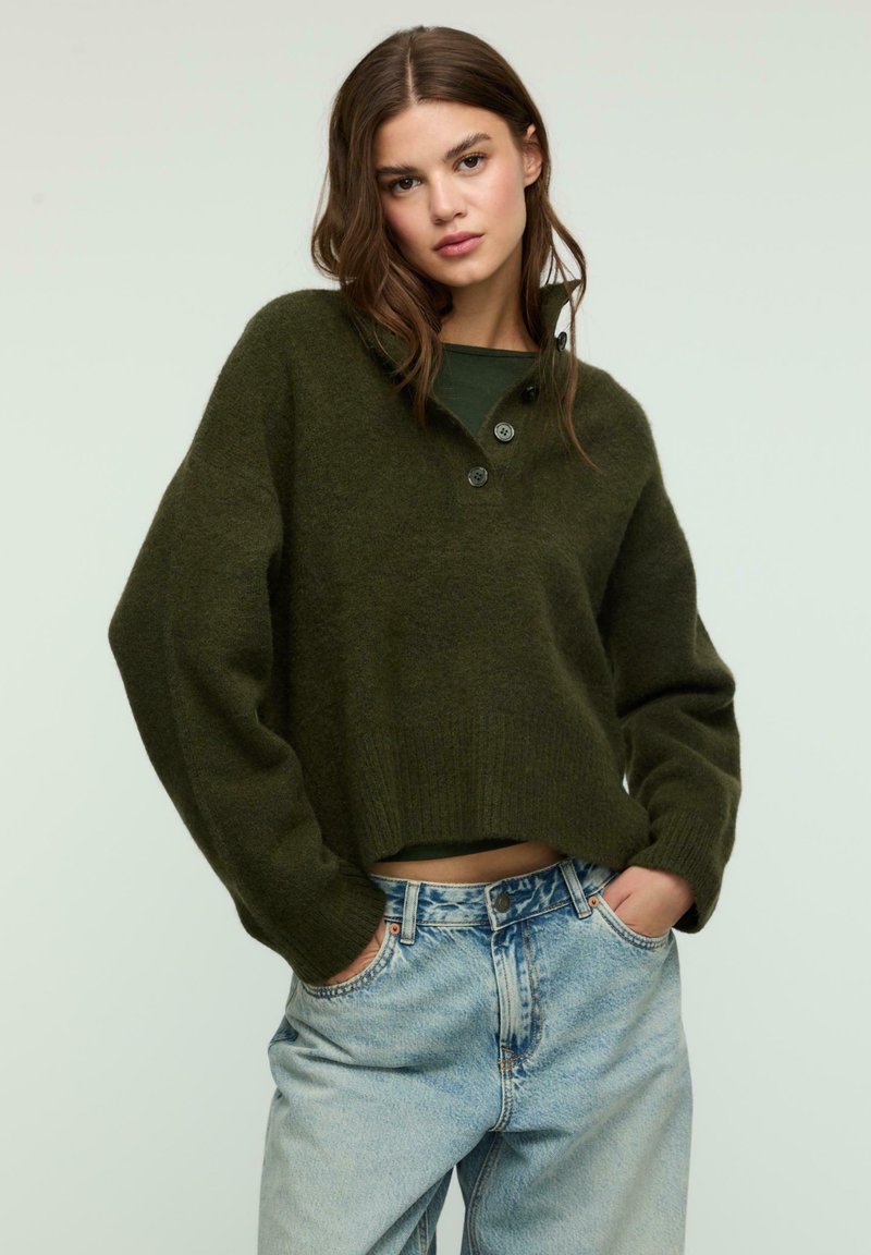 Maglione corto verde oliva con colletto, tre bottoni e polsini a costine, abbinato a jeans alta vita azzurri. Sfondo neutro.