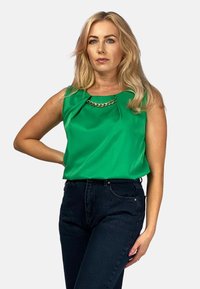 Zielona, bezrękawnikowa bluzka z plisowanym dekoltem i z łotym akcentem w postaci łańcuszka; zestawiona z ciemnymi, wysokim stanem dżinsami, co tworzy kontrastowy wygląd.