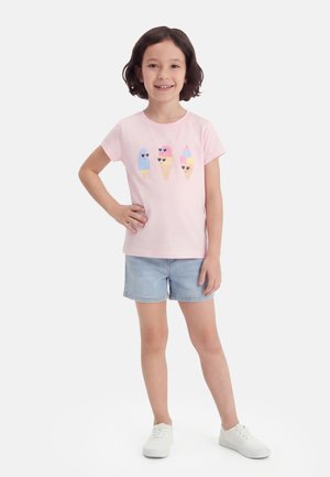 Fille debout, main sur la hanche, portant un t-shirt rose clair avec des motifs de glace, un short en jean clair et des baskets blanches.