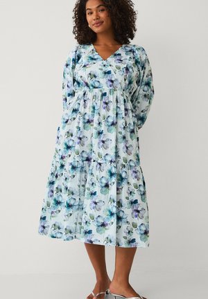 Vrouw met krullend haar die een midi bloemenjurk met lange mouwen draagt met blauwe en groene bloemen en zilveren open tenen hakken, staand tegen een effen achtergrond.