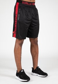 Zwarte sportshorts met rode accenten en logo, voorzien van een verstelbare tailleband en een ritszak aan de zijkant. Het materiaal lijkt lichtgewicht en glad te zijn.