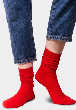 Czerwone, prążkowane skarpetki z marszczonymi ściągaczami, pokazane na nogach w krótkich niebieskich dżinsach. Skarpetki mają gładką teksturę i jasny kolor.