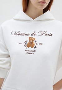 Witte hoodie met geborduurde tekst "Avenue de Paris", met een ontwerp van een bruine beer en decoratieve bloemdetails.