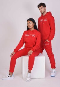Conjunto de sudadera con capucha y pantalones de chándal a juego en rojo, con un logo blanco y cordón. Ambos modelos llevan zapatillas deportivas. Textura de tela suave.