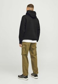 Moški obrnjeno stran, oblečen v črno kapuco, belo srajco, olivno zelene hlače s žepi in črne superge na svetlo sivi podlagi.