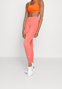 Reggiseno sportivo Nike di un vivace arancione e leggings rosa a vita alta, entrambi aderenti. Logo Nike bianco sui leggings, abbinati a scarpe da ginnastica bianche.