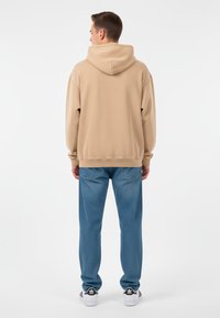 Man som står vänd bort, iförd en beige hoodie, blå jeans och vita Adidas-sneakers mot en vit bakgrund.