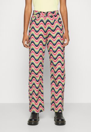 Person iført højtaljede bukser med lige ben og flerfarvede bølgede striber i pink, blå, gul og sort, kombineret med sorte støvler.