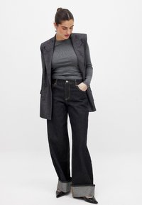 Chaleco gris a medida sobre una camiseta gris de canalé, combinado con jeans de mezclilla oscuros de pierna ancha con puños doblados y tacones negros puntiagudos.
