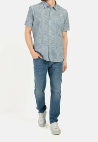 Camisa de botones de manga corta en azul con una textura estampada, combinada con unos jeans azul claro y zapatillas blancas. El modelo está de pie con las manos en los bolsillos.