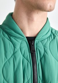 Zbliżenie osoby noszącej zieloną pikowaną kurtkę z czarnym zamkiem błyskawicznym, ukazujące szyję i dolną część twarzy z profilu.