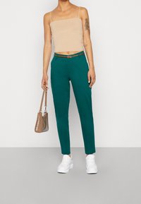 Tyrkysové kalhoty s úzkým střihem, hnědý pásek, spárované s béžovým topem. Modelka drží malou hnědou kabelku a má na sobě bílé tenisky.