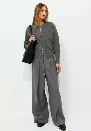 Mujer vestida con un cárdigan gris abotonado, pantalones grises anchos, zapatos negros y llevando un bolso negro sobre el hombro.
