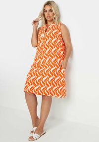 Oranssi hihaton mekko, jossa on valkoinen liekkikuosi, napitettava kaula-aukko ja sivutaskut. Yhdistetty valkoisiin sandaaleihin.