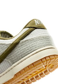 Hvid og olivengrøn Nike-sneaker tæt på, der viser tekstureret stofoverdel, olivengrøn kant, logo på hælen og slidmønster på gummiydersålen.