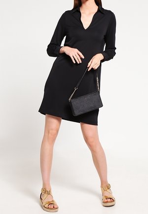 Donna che indossa un vestito nero a maniche lunghe con una piccola borsa nera, abbinata a sandali dorati con plateau e cinturini.