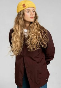 Czerwono-bordowa kurtka o błyszczącej powierzchni z zatrzaskami, bocznymi kieszeniami i elastycznymi mankietami. Noszona z żółtą dzianą czapką o teksturowanej powierzchni.