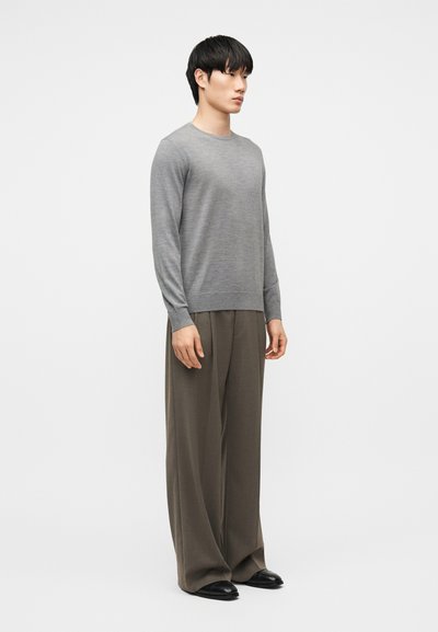 Muž stojící v šedém tričku s dlouhým rukávem, volných hnědých kalhotách a černých botách na světle jednobarevném pozadí.