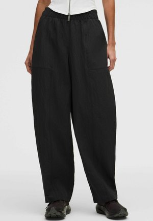 Lockere schwarze Hose mit elastischem Bund, getragen mit schwarzen Schuhen und weißem Reißverschluss-Oberteil, gezeigt von der Taille bis zu den Füßen vor hellem Hintergrund.