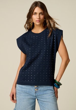 Femme aux longs cheveux bruns porte un débardeur bleu marine avec des petits clous argentés et un jean bleu clair, accessoirisée avec des bracelets colorés.