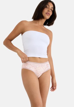 Femeie purtând un top scurt alb fără bretele și lenjerie intimă roz deschis cu imprimeu floral, stând în fața unui fundal simplu de culoare deschisă.